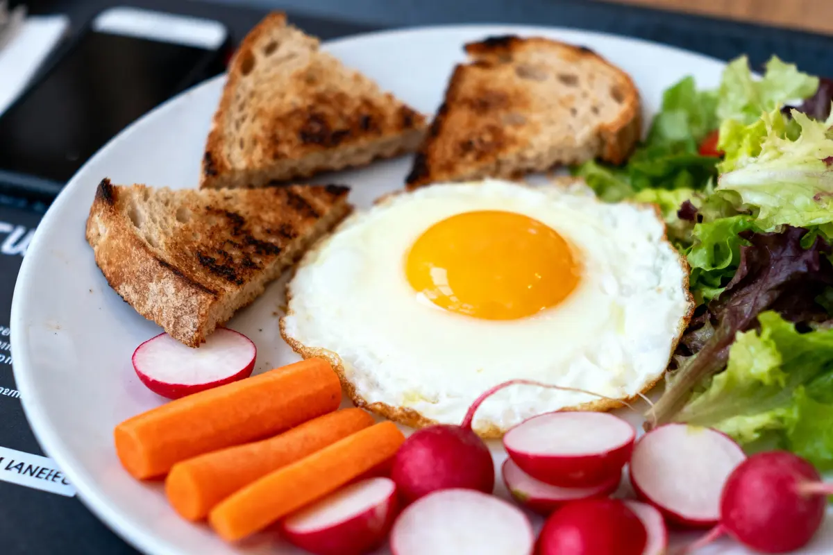Assiette contenant œuf au plat, toasts grillés, radis, carottes et salade verte variée sur fond sombre.