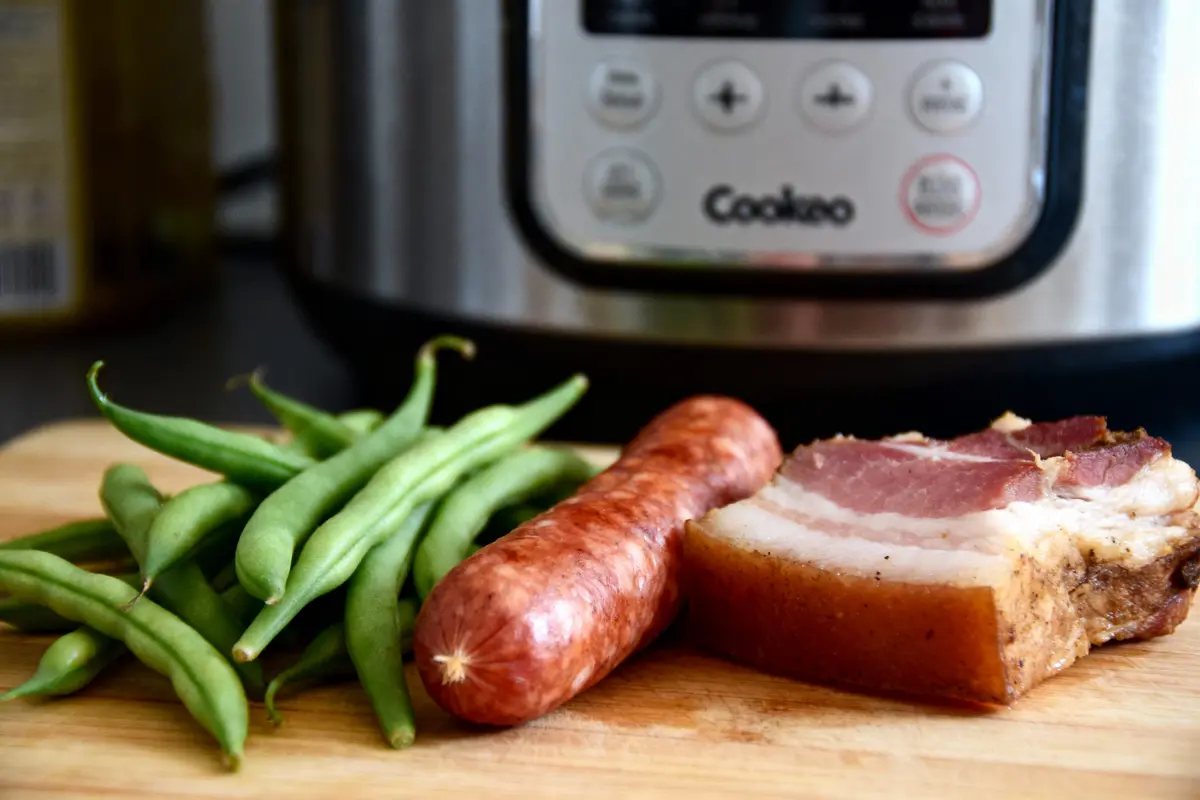 Haricots verts, saucisse et lardons sur planche devant un autocuiseur argenté.