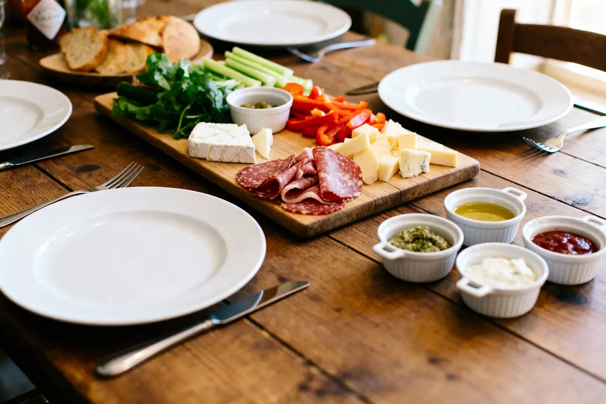 Planche de charcuterie avec fromages, légumes et condiments sur table en bois, assiettes blanches et couverts alentours.