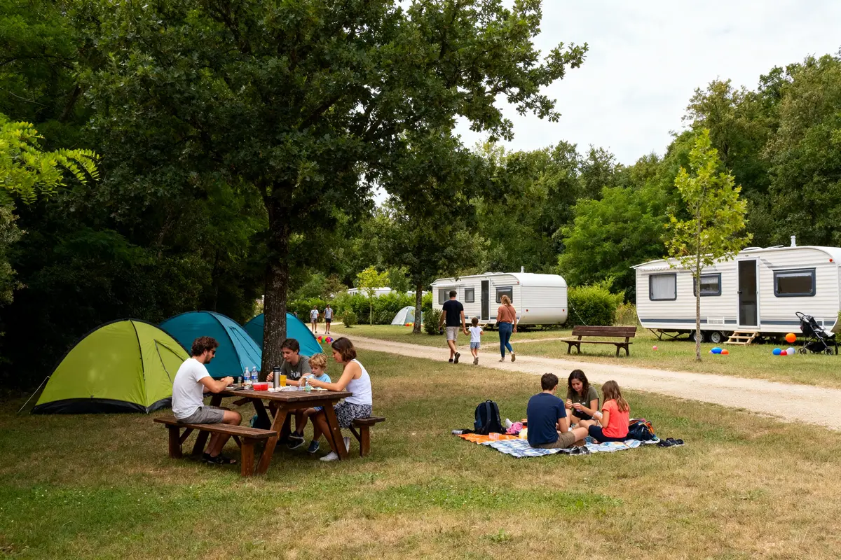 Groupe de personnes en camping, avec tentes, tables, caravanes et arbres environnants, profitant de la nature.