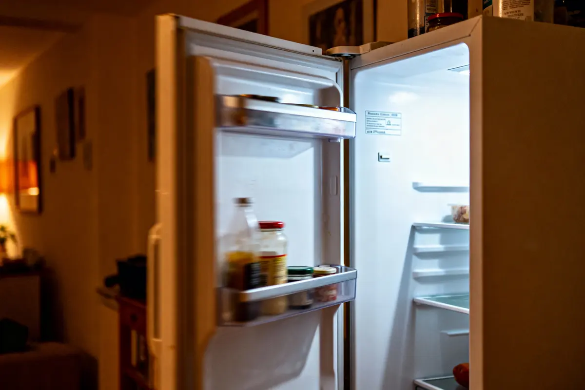 Réfrigérateur ouvert dans une cuisine sombre, lumière intérieure illuminant bocaux sur la porte et étagères vides.