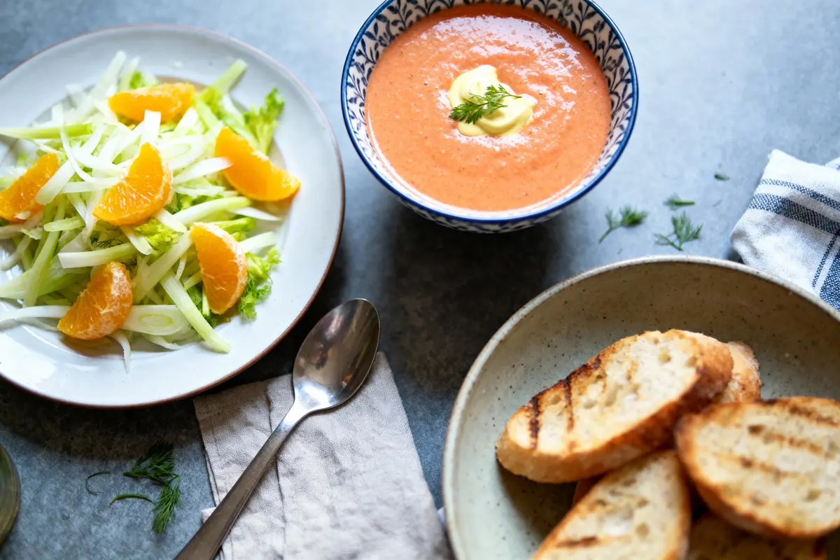 Assiette de salade fenouil-orange, soupe tomate-crème, pain grillé sur table grise avec serviette blanche.