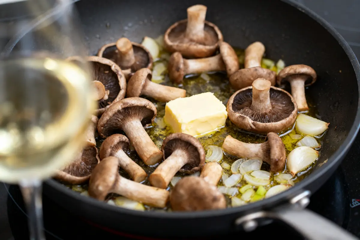 Champignons cuisinés dans une poêle avec beurre, oignons, et fond de verre de vin blanc visible.