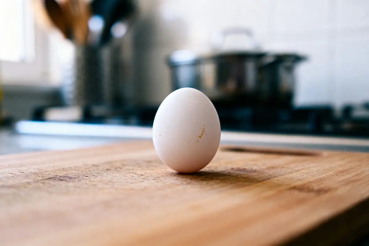 Un œuf blanc posé sur une planche en bois dans une cuisine moderne en arrière-plan.