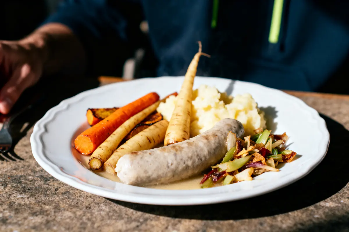 Assiette avec saucisse, purée de pommes de terre, légumes rôtis, sur une table en bois, éclairage naturel.