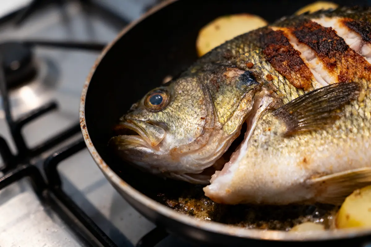 Poisson entier frit dans une poêle sur une cuisinière, avec pommes de terre en rondelles.