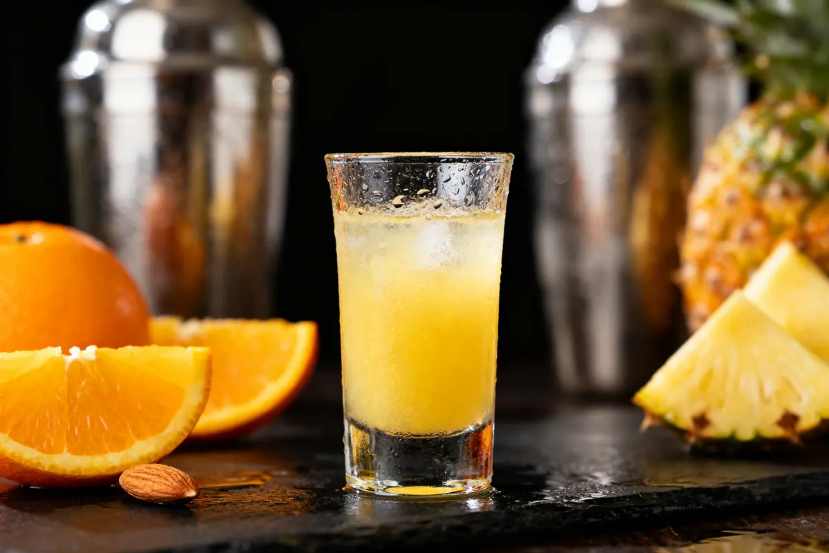 Verre de cocktail jaune avec glaçons, entouré d'orange et ananas, devant des shakers en métal.