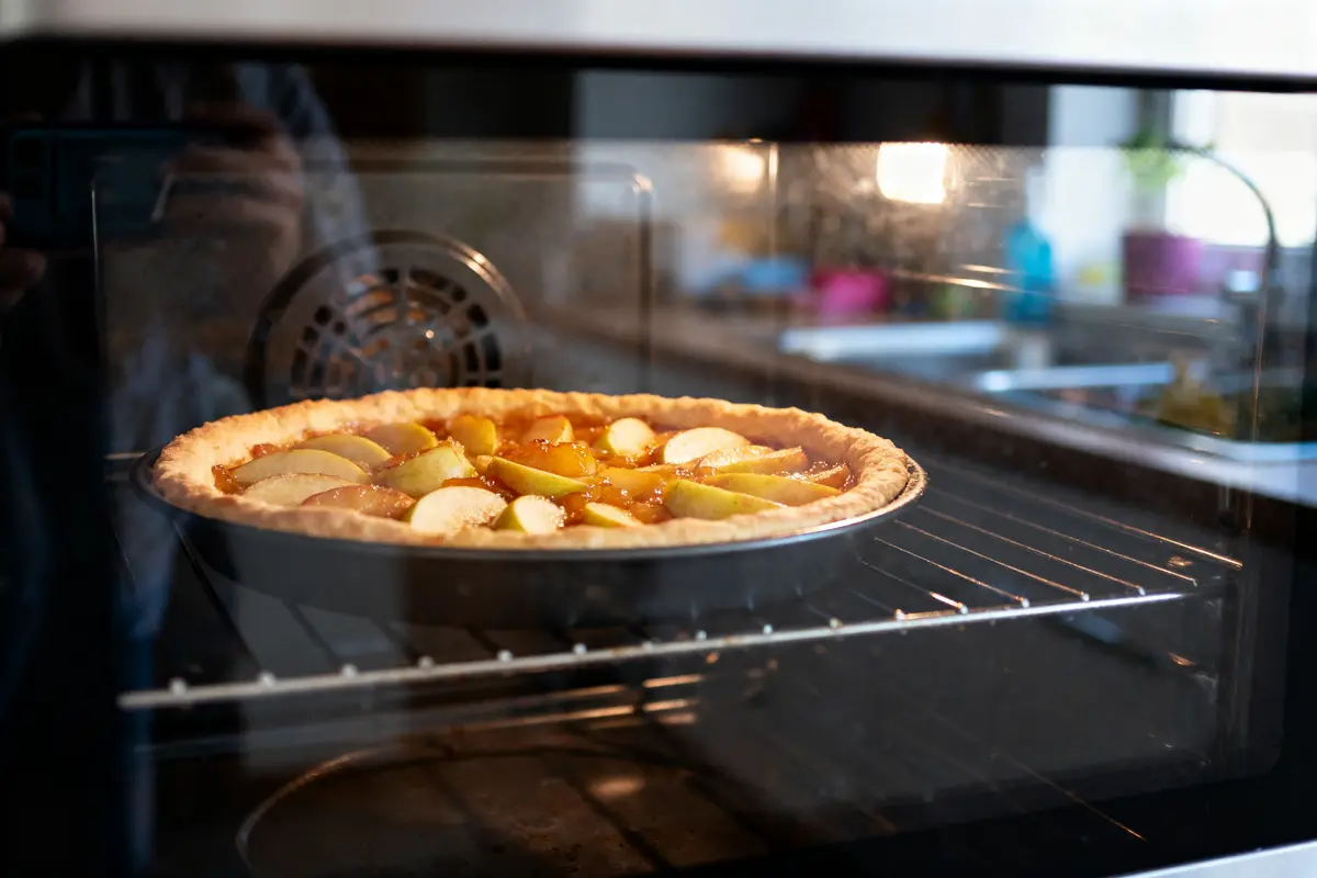 Tarte aux pommes cuisant dans un four, croûte dorée et tranches de pommes visibles, cuisine en arrière-plan.